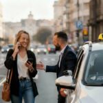 Putnica razgovara telefonom i drži drugi uređaj dok taksista otvara vrata pored belog taksija sa natpisom TAXI, gradska ulica u pozadini.